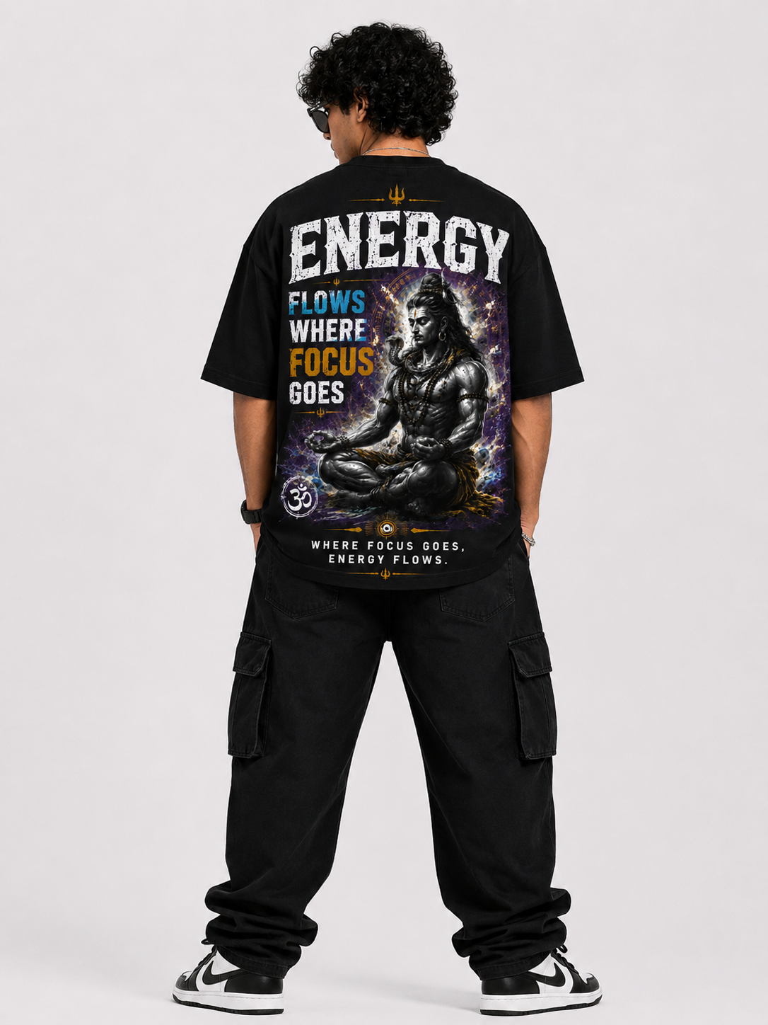 A man viewed from behind wearing a black graphic t-shirt with a large back print featuring a seated figure and the text 'ENERGY FLOWS WHERE FOCUS GOES'.