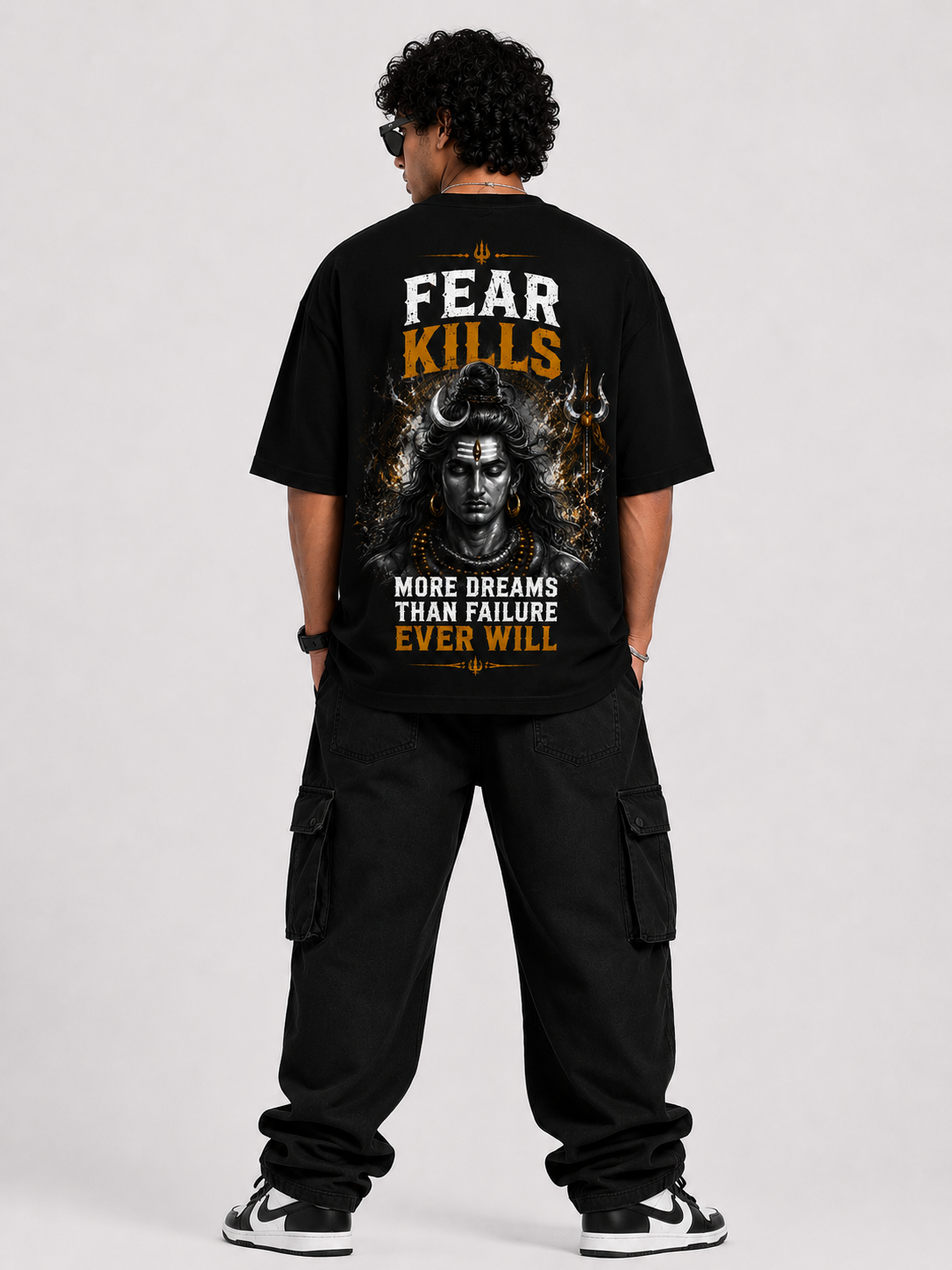 A man viewed from the back wearing a black graphic t-shirt with a large design featuring the text 'FEAR KILLS' and 'MORE DREAMS THAN FAILURE EVER WILL' alongside a stylized face illustration, paired with black cargo pants and black-and-white sneakers, against a light gray studio background.