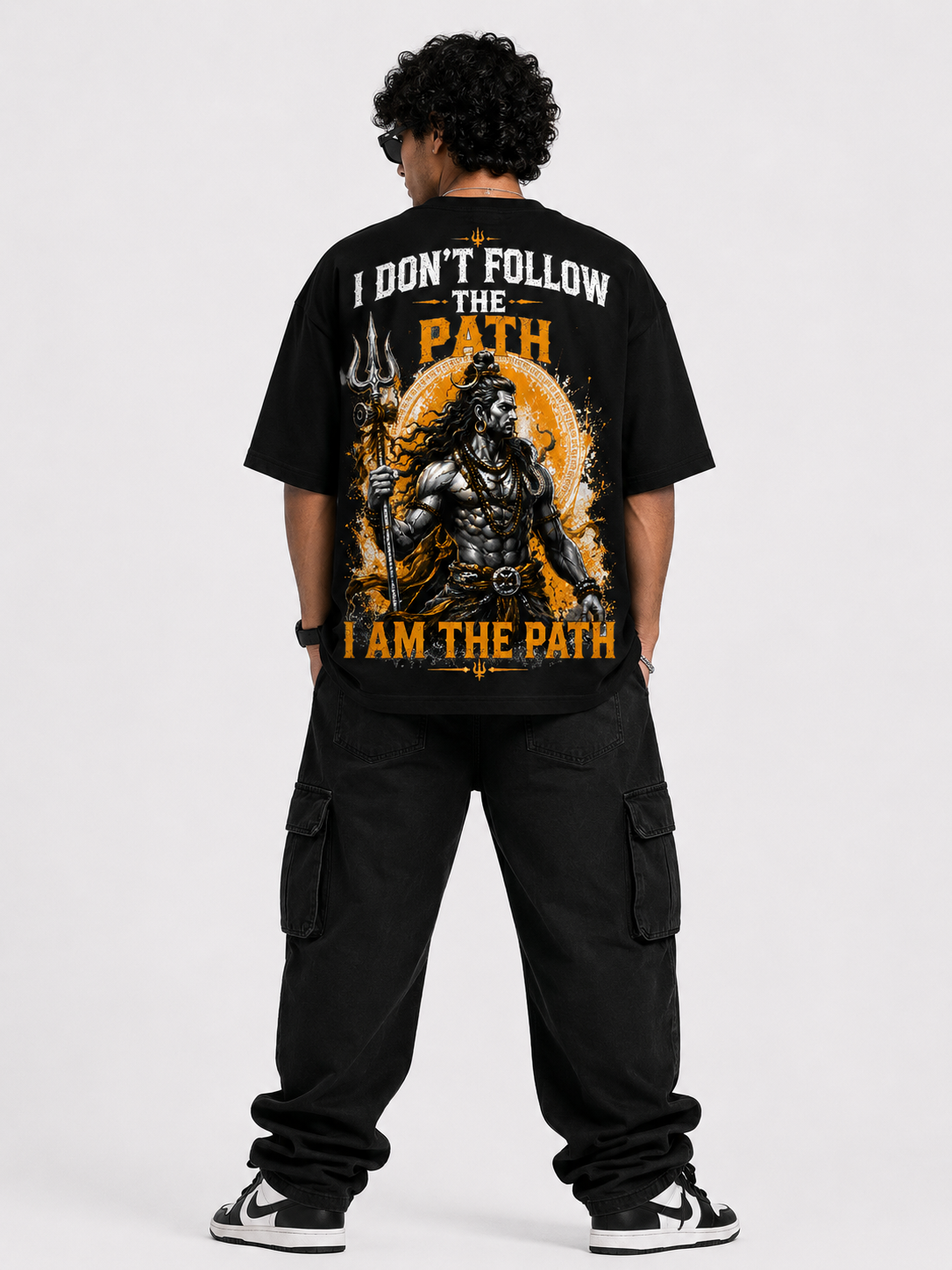 A man viewed from the back wearing a black graphic t-shirt with a large print of Lord Shiva and the text 'I DON'T FOLLOW THE PATH I AM THE PATH', paired with black cargo pants and black and white sneakers, against a light gray studio background.