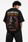 A man wearing a black oversized graphic T-shirt, shown from the back. The shirt features a large print of a seated figure with a trident on a purple and orange cosmic background, with the text 'THE MIND QUITS THE SOUL WINS' and 'YOU ARE STRONGER THAN YOU THINK'.