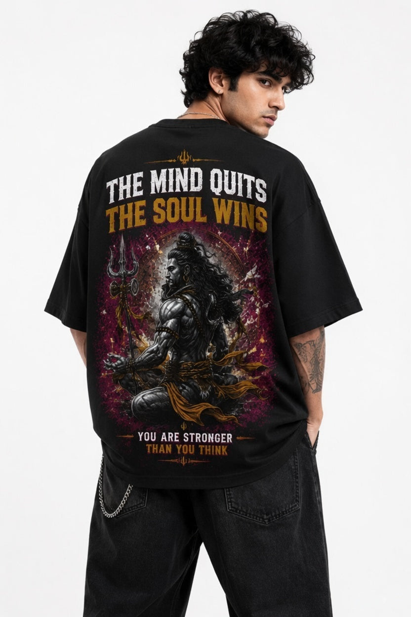 A man wearing a black oversized graphic T-shirt, shown from the back. The shirt features a large print of a seated figure with a trident on a purple and orange cosmic background, with the text 'THE MIND QUITS THE SOUL WINS' and 'YOU ARE STRONGER THAN YOU THINK'.