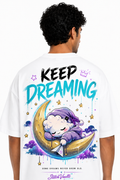 A man viewed from behind wearing a white t-shirt with a colorful graphic on the back. The graphic features the text 'KEEP DREAMING' in black and teal, a cartoon character sleeping on a crescent moon, and the brand name 'SketchVaults' at the bottom.
